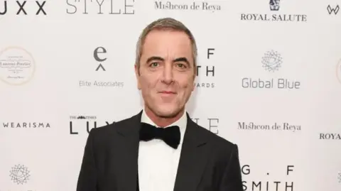 Getty Images A man with dark grey hair is smiling and looking toward the camera. He is dress in a formal black suit with a black bow tie and a white shirt. There is branding on the wall behind him. 