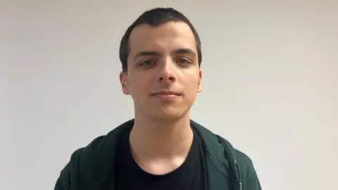 Young man with black hair and a black-tshirt. He is smiling at the camera.