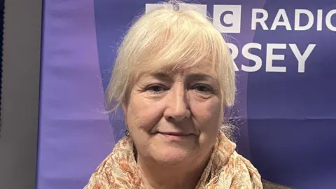 BBC Deputy Karen Wilson is standing in front of a BBC Jersey banner. She is wearing a brown blazer and a floral scarf. 