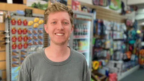 A man stands at the front of a gift shop in a seaside town in Cornwall. He is wearing a t shirt and there are buckets, spades and body boards on the walls behind him.