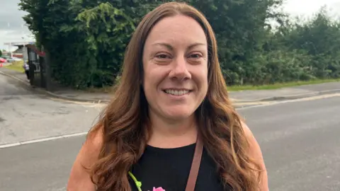 Sarah Foster. She is pictured outside. She has long brown hair and is looking straight at the camera and smiling.