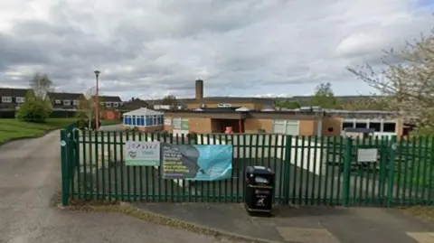 The entrance to Westfield Infant School, which is behind a tall green metal fence. A paved path leads through the gate and curves to the left, bordered by grass and trees.
Beyond the fence, the buildings are low and made of light brown brick, with flat roofs and several windows and doors. Banners and signs are attached to the fence, and a black litter bin stands near the entrance.