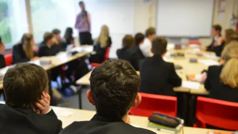 A classroom with the backs of the heads of two boys in the foreground with other out of focus pupils sitting around desks and a teacher standing at the front of the class