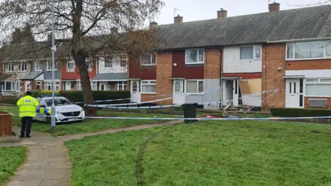 A terraced house cordoned by police tape with the front door and window severely damaged. A police officer is outside the house, standing next to a police vehicle.