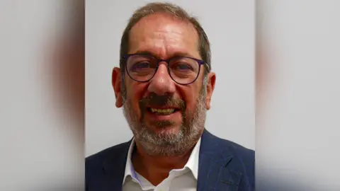 Broudie Jackson Canter Close up of Elkan Abrahamson with brown hair and beard wearing brown glasses and a navy suit with a white shirt. He is pictured in front of a light grey background. He is smiling.