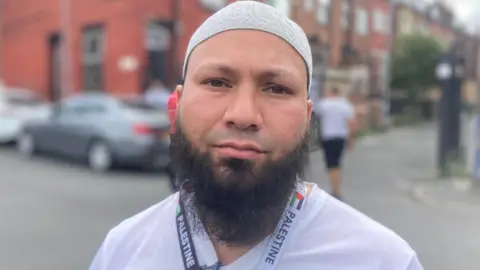 A man with dark eyes and a dark beard is wearing a grey skull cap, white T-shirt and a lanyard with the word Palestine written on it. Behind him is a blurred view of a red brick building, a silver car parked on a road and a man walking away.
