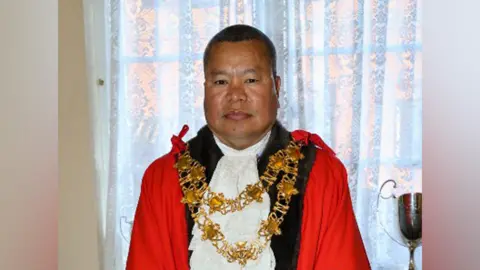 A nepalese man in a red mayor's cloak and white crevette, as well as gold mayoral chains. He has a shaved head and is smiling. He is standing in front of a white net curtain. 