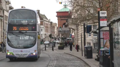 A purple and white First bus driving down Colchester High Street.
