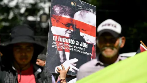 Orlando SIERRA / AFP via Getty Images) A woman holds a banner reading "The pardon for Juan Orlando Hernandez (JOH) does not erase the truth, does not erase the narco-state", during a protest outside the US Embassy in Tegucigalpa on 4 December, 2025