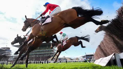 PA Media Three horses land after jumping fences. They are seen from below and are being ridden by jockeys during the Gold Cup race at Cheltenham. Large stands are in the distance. 