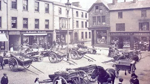 A black-and-white photo from the early 1900s depicting several horse carts resting on a cobbled pavement while people mill about. Goods are being sold in the carts. There are old Victorian shop fronts in the background. 