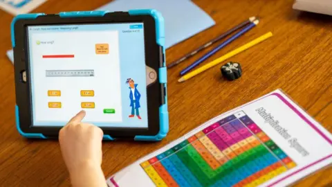 A child's hand reaching out to use an iPad while a homework sheet sits next to it. The rest of the child is not in the shot.