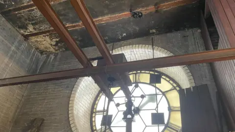 Simon Thake Wooden beams criss-cross a room with stone walls. The back of a large clock face dominates the image