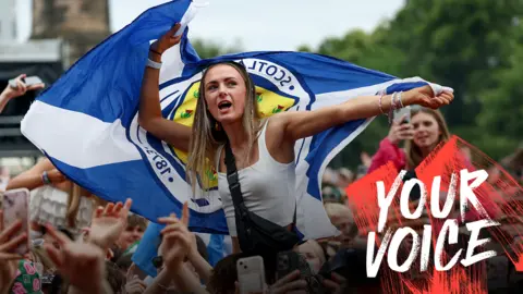 A young woman in a white sleeveless tops is sitting on someone's shoulders, above a large crowd of people. She is waving a Scotland flag. 
