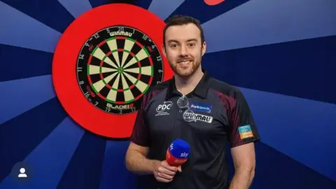 Huw Ware smiles looking at the camera. He is standing in front of a dart board and is holding a mic.