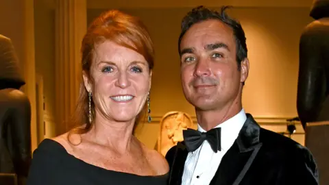 Getty Images Sarah Ferguson in black dress with her hair up stands next to Manuel Fernandez who wears a black velvet jacket, white shirt and bow tie. They are both smiling at the camera. 