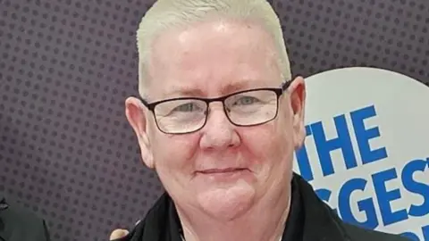 North West Pest and Rodent Control Patricia Page with short white hair stands in front of an interior wall. She is wearing a smile and glasses, and a black jacket.