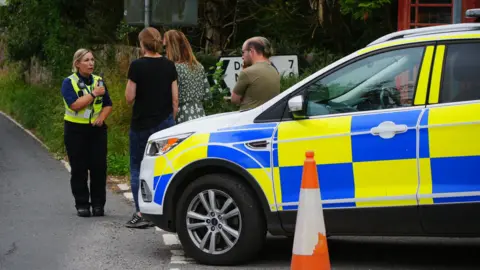 A female police officer in uniform talks to local residents standing next to a police car in Wheddon Cross in Somerset after a coach crash