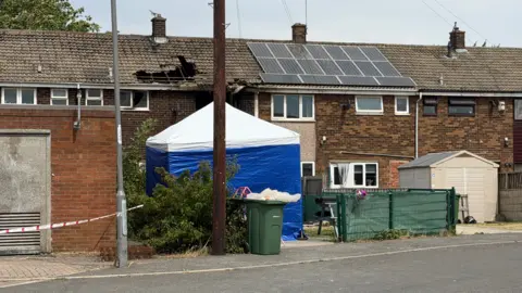 A blue and white police tent at the scene. A hole has been burnt into the roof of a terrace house.