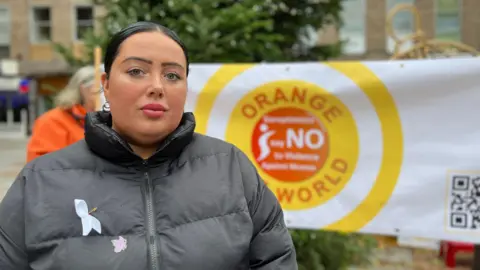 Suzi looks at the camera as a sign is held up behind her saying say no to violence against women and organe is the world. Suzi is wearing a black coat without a hood and has a white ribbon pinned to her chest. She has black hair which is short and tied into a pony tail.