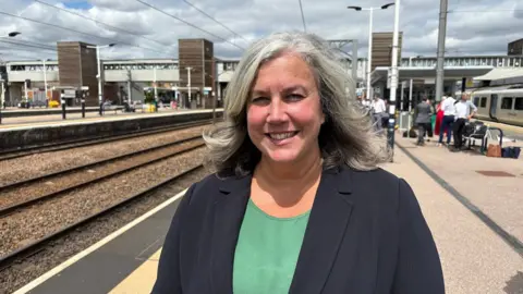 Heidi Alexander smiles at the camera, dressed in a green shirt and black jacket. She is standing outside on a platform at a train station. There are a number of people in the background. 