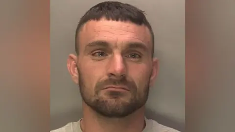 A police mugshot shows a man with brown hair and stubble. He is wearing a grey t-shirt.