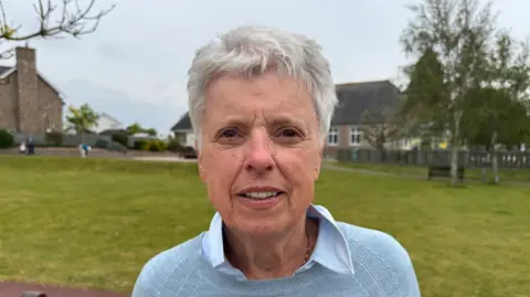 Olive Thomas is wearing a blue jumper and blue shirt, she is pictured in St Ouen's village