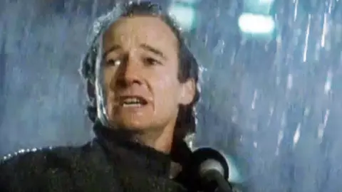 David Hayman in 1986, with long hair and in an overcoat, is standing in heavy rain speaking into a microphone. He looks passionate and serious.