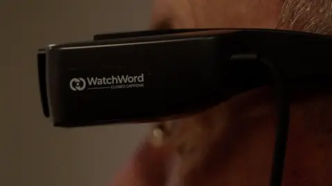 The side of a pair of black smart glasses with "WatchWord Closed Captions" inscribed on the side in white lettering. You can see a bit of grey hair and an out of focus man's face.