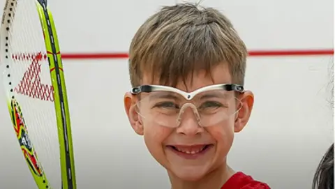 An eight-year-old boy with short brown hair and a fringe, smiling towards the camera, wearing clear, safety glasses and holding a squash racquet. 