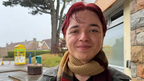 Toby Cahill Le Broq is pictured in St John, he has red hair and a yellow, brown and red scarf. 