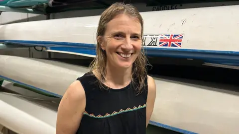 A woman with long dark blonde wet hair, wearing a black sleeveless top, stands in front of several kayaks lying on racks.