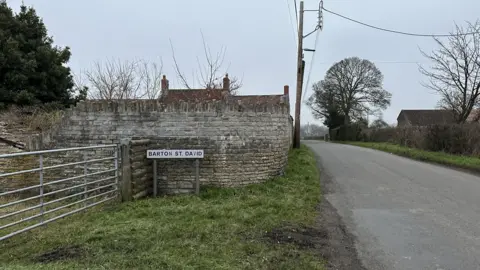 A road sign reading 'Barton St David' at the side of the road. There is a wall behind it and a fence next to it. There's a power line in the sky and the sign is on a small patch of grass.