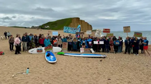 Surfers Against Sewage A group of sign waving protestors on a beach with paddleboards at their feet.