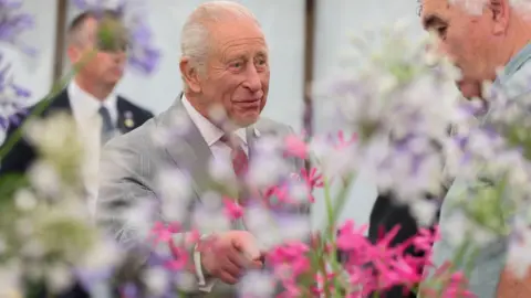  King Charles III during a visit to the Sandringham Flower Show. He is in a marquee and standing before some pink nerines and pointing them to another man.  