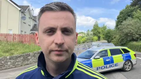 A man in a sports top is standing at a police cordon. The police car is blue, white and yellow.