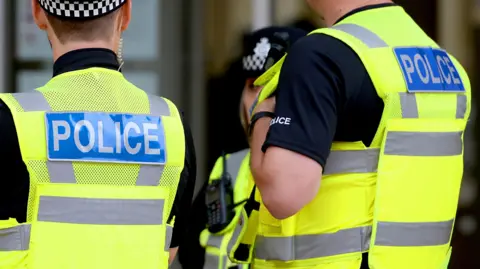 An image of three police officers who are not connected to the incident with their backs to the camera. They are wearing high viz vests with police written on their backs.