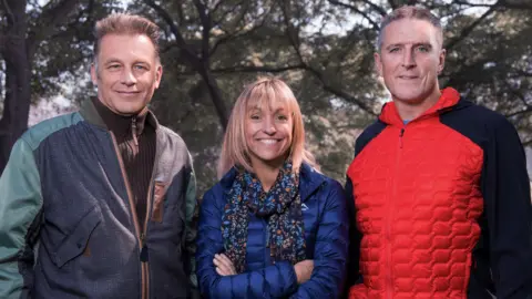 Chris Packham, Michaela Strachan and Iolo Williams stand in a row in front of trees and smile at the camera. On the right, Chris Packham, who has short, light coloured hair, is wearing a brown ribbed jumper and a patchwork zip-up jacket. In the middle, Michaela has blonde, medium-length hair with a full fringe and is wearing a blue puffed jacket and a blue and orange patterned scarf. She has her arms crossed across her chest. On the right, Iolo has short grey hair and is wearing a red and black padded zip up raincoat.