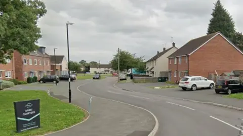A Google street view image showing a curved, residential street with houses on either side and cars parked in driveways.