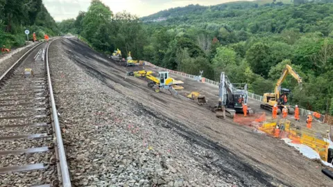 A section of railway track running to the left hand side of the photograph, with a sloping downwards hill to the right and workmers in orange high vis with diggers at work. 