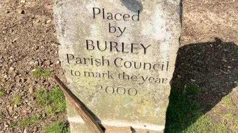 A memorial stone bearing the words "Placed by Burley Parish Council to mark the year 2000", with an upside down wooden cross placed at its foot 