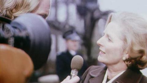 Margaret Thatcher on right, being interviewed outside by a journalist on the left who is obscured by a camera lens, holding a microphone in front of her mouth.  In the background a policeman is standing in front of a row of houses.
