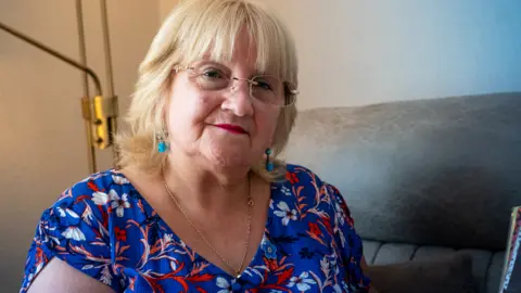 Yeti Television/BBC Cymru Wales Pauline Watkins is a white woman in late middle age. She has dyed blonde shoulder-length hair with a fringe. She wears a V-necked royal blue dress with a pattern of large white, red and black flowers on it and a gold chain with an unseen locked underneath the dress. She wears rimless glasses and turquoise drop earrings. She wears blue eye shadow and red lipstick and is looking directly at the camera with a very slight smile. She is seated on a sofa in a living room, with standard light gold poles behind her.