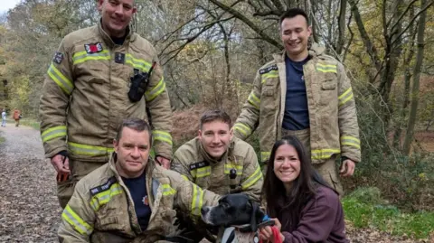 Essex Fire and Rescue Four firefighters, a dog and a woman in a wooded area. Two firemen are standing, and two are crouching next to a black and white dog and its owner, a woman wearing a purple jacket