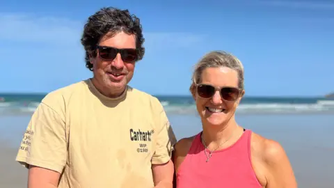 Nick and Rebecca are wearing sunglasses stand together on a wide sandy beach. The ocean and a clear blue sky stretch behind them, with the shoreline visible in the distance.