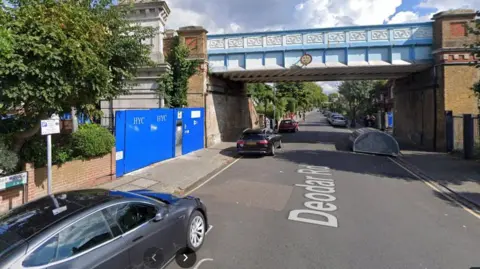 A Google streetview screengrab of Deodar Road, there is a grey car to the left, parked, and a blue railway bridge ahead, as well as a bike rack to the right on the road. 