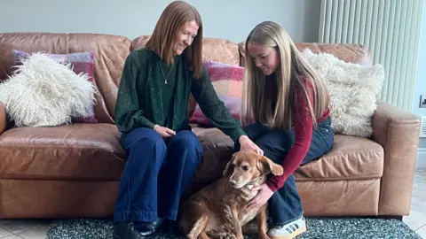Emily and her mum Angela sitting on a brown leather sofa stroking their dog. The dog is brown short haired. The sofa has a mixture of fluffy cream cushions and patterned ones on it.