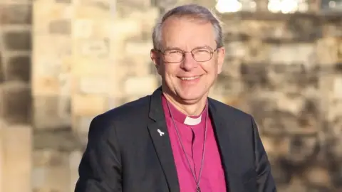 Church of England The Right Reverend Paul Butler wearing a reverend's collar and black blazer. He is smiling and wears glasses. He has a purple top on and a chain. 