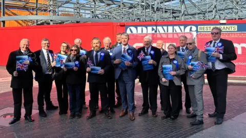 Local Democracy Reporting Service A large group of Conservative Party candidates posing outside the redevelopment of the western end of Freshney Place. There are women and men and they are all wearing either suits or office wear. They all have badges on and are holding British flags and blue leaflets. 