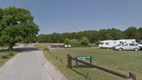 A wide view of Roundhill campsite with an oak tree beside a narrow road and several cars and caravans parked up on an area of grass to the right.